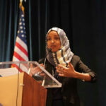 Omar VANISHES Before $250M Fraud Testimony… Woman speaking at podium with American flag background.