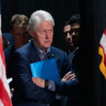 Bill Clinton CRASHES America’s Team Press Conference… Bill Clinton standing between American flags with a folder.