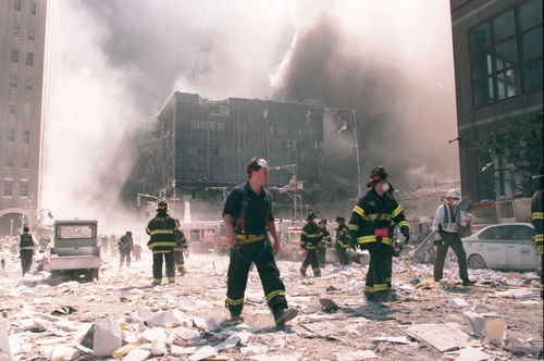 Firefighters walking through debris near a heavily damaged smoking building