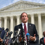 Residency Dispute puts on a Political Collision Course Men in suits speaking at podium outside courthouse.