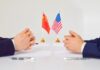 Two hands of individuals facing each other with flags of China and the USA on the table
